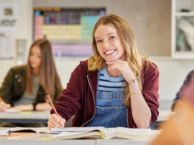 Eine junge Frau sitzt im Vordergrund an einem Tisch in einem Klassenzimmer oder Seminarraum. Sie trägt eine dunkelrote Jacke über einem blau-weiß gestreiften Oberteil und einem blauen Overall. In der rechten Hand hält sie einen roten Stift und stützt ihr Kinn nachdenklich auf die linke Hand. Vor ihr liegen ein offenes Notizbuch und weitere Schulmaterialien. Im Hintergrund sind weitere Personen zu erkennen, die ebenfalls an Tischen sitzen und arbeiten. An der Wand hängt eine bunte Pinnwand mit verschiedenen Notizen und Materialien. Die Atmosphäre wirkt konzentriert und lernorientiert.
