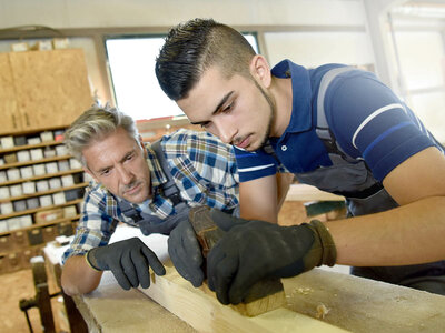 Zwei Personen arbeiten an einem Stück Holz in einer Holzwerkstatt. Beide tragen Handschuhe und benutzen Werkzeuge, um das Holz zu glätten oder zu formen. Im Hintergrund sind Regale mit verschiedenen Gegenständen zu sehen, die wahrscheinlich mit der Holzbearbeitung zu tun haben.
