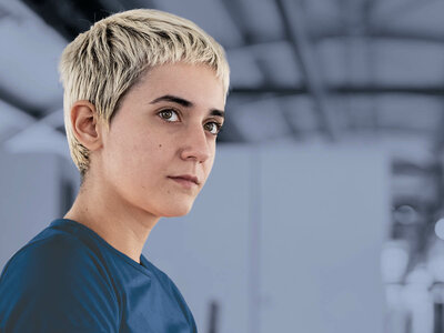 Eine Person mit kurzen, blonden Haaren und einem blauen T-Shirt steht in einem Fitnessstudio oder einer Sporthalle. Die Person blickt nachdenklich oder konzentriert zur Seite, während der Hintergrund leicht unscharf ist.