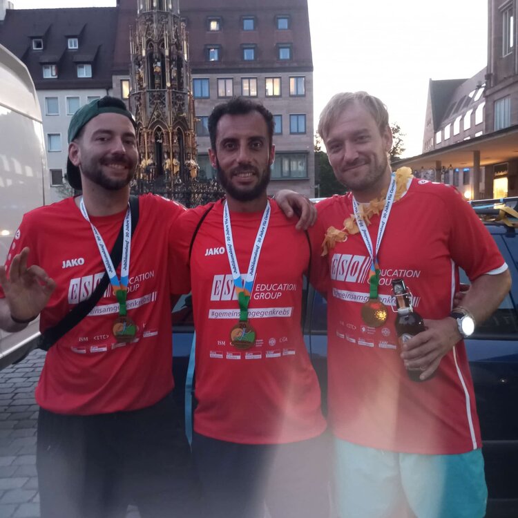 Drei Teilnehmer des Benefizlaufs stehen nach dem Zieleinlauf mit Medaillen in roten ESO-Trikots vor dem Schönen Brunnen in Nürnberg.