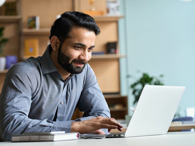 Ein lächelnder Mann mit Bart und einem grauen Hemd sitzt an einem Schreibtisch und arbeitet konzentriert an einem Laptop. Auf dem Tisch liegen ein Notizbuch und ein Stift. Der Mann wirkt entspannt und zufrieden. Im Hintergrund sind Regale mit Büchern und Ordnern sowie eine Zimmerpflanze zu sehen, was auf ein Büro oder Homeoffice hindeutet. Der Raum wirkt freundlich und gut beleuchtet.