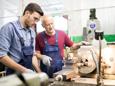 Zwei Personen stehen in einer Werkstatt oder Fabrikumgebung neben einer Drehmaschine, die gerade ein Metallstück bearbeitet. Beide tragen Overalls und Handschuhe. Im Hintergrund sind verschiedene industrielle Geräte und Werkzeuge zu sehen.
