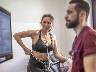 Das Bild zeigt eine kardiovaskuläre Funktionsdiagnostik, bei der eine Frau mit Überwachungsgeräten während körperlicher Aktivität verbunden ist, um die Herzgesundheit in Echtzeit zu beurteilen. Ein Mann überwacht die Werte an einem Monitor im Vordergrund.