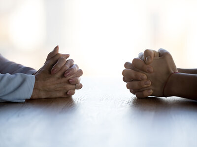 Zwei Personen sitzen sich an einem Tisch gegenüber und haben ihre Hände gefaltet. Die Hände sind im Vordergrund zu sehen, während die Gesichter der Personen nicht sichtbar sind. Die Szene wirkt ruhig und nachdenklich, möglicherweise in einem Gespräch oder einer Besprechung.
