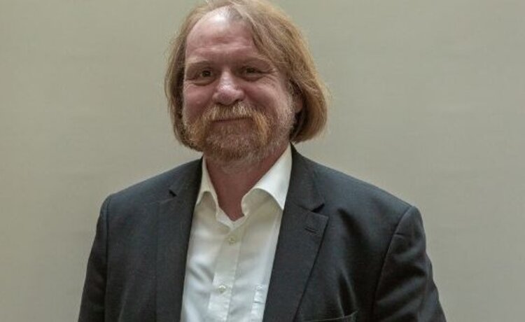 Porträtfoto von Volker Hiekel, der einen dunkelgrauen Herrenblazer und ein weißes Hemd sowie ein Lächeln auf den Lippen und einen Bart trägt.