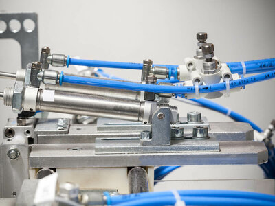 Nahaufnahme einer mechanischen Baugruppe mit mehreren blauen Pneumatikschläuchen, die an Metallkomponenten angeschlossen sind. Es gibt verschiedene Anschlüsse, Bolzen und einen Zylinder, die Teil einer Industriemaschine oder eines Automatisierungssystems zu sein scheinen. Die Schläuche sind mit Text beschriftet, der jedoch im Bild nicht vollständig lesbar ist.