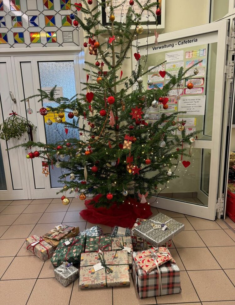 Ein geschmückter Weihnachtsbaum steht in einer Eingangshalle vor der Tür der Kolpingschule in Aschaffenburg. Die Tür hat die Beschriftung: "Verwaltung - Cafeteria". Der Baum ist mit roten und goldenen Kugeln, Herzschmuck, Lametta und Lichterketten dekoriert. Um den Baum herum liegen zahlreiche bunt verpackte Geschenkpäckchen in verschiedenen Größen und Mustern auf dem Boden. Im Hintergrund sind farbenfrohe Glasmosaiken zu sehen.