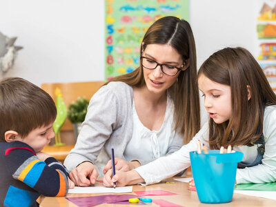 Auf dem Bild sitzen zwei Kinder und ein Erwachsener an einem Tisch und zeichnen. Bunte Zeichnungen und Lernmaterialien schmücken die Wände.