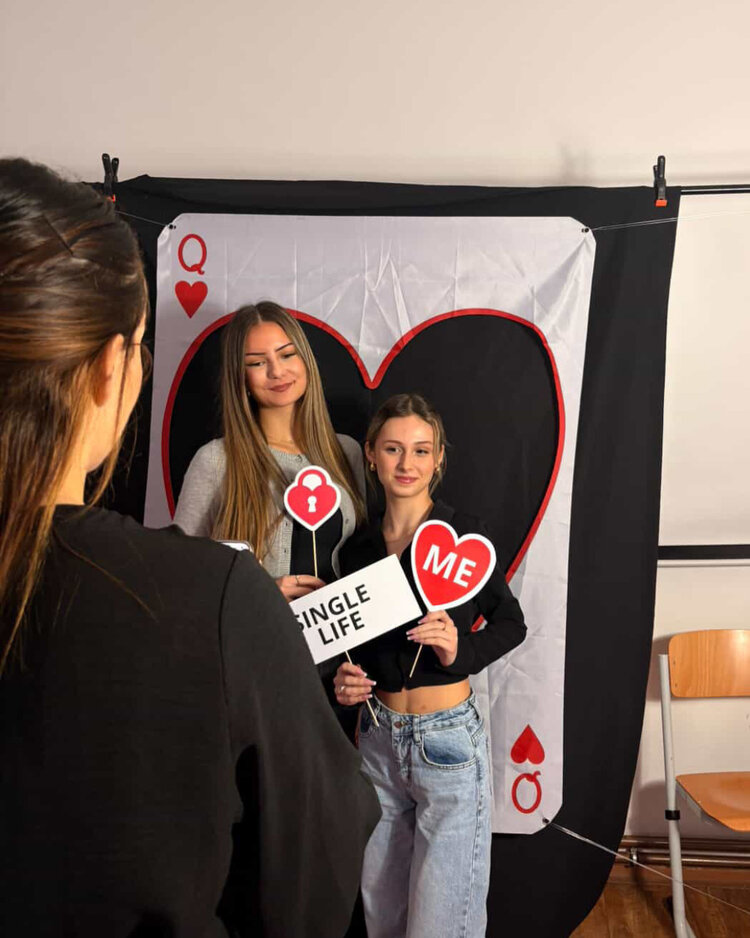 Zwei junge Frauen posieren vor einem großen Spielkartenmotiv (Herz Queen). Eine hält ein Schild mit der Aufschrift "Single Life", die andere ein Schild mit einem Herz und der Aufschrift "ME". Eine Person im Vordergrund fotografiert sie.
