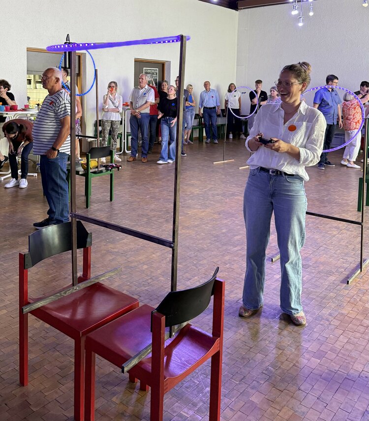 Eine Frau steuert lachend eine Drohne durch einen beleuchteten Parcours aus Ringen und Gestellen, der in einer Halle aufgebaut ist. Um sie herum stehen und beobachten viele Menschen das Geschehen. Der Raum wirkt wie eine Veranstaltungs- oder Mehrzweckhalle mit Holzboden.