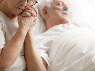 Auf dem Bild sieht man eine ältere Frau, die die Hand ihres Mannes am Krankenbett hält.