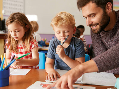 Das Bild zeigt zwei Kinder und einen Erwachsenen, die an einem Tisch sitzen und zeichnen oder schreiben. Auf dem Tisch liegen Papiere, und im Hintergrund ist eine Klassenzimmereinstellung mit Bildungsmaterialien an den Wänden zu sehen.