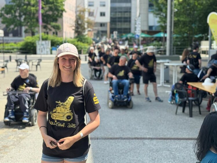 Eine junge Frau mit hellblonder Haaren, schwarzem T-Shirt mit der Aufschrift "Rollitour 2025" und einer beigefarbenen Kappe steht lächelnd im Vordergrund. Im Hintergrund sieht man zahlreiche Menschen in Rollstühlen und zu Fuß, die gemeinsam an der Rollitour teilnehmen. Die Veranstaltung findet bei sonnigem Wetter auf einem städtischen Platz statt.