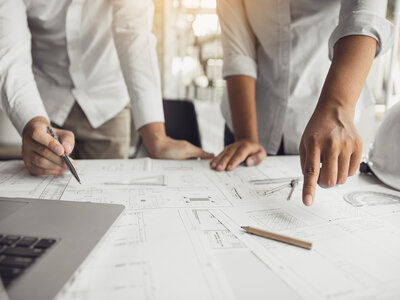 Eine Gruppe von Personen arbeitet an einem Tisch mit verschiedenen technischen Zeichnungen und Plänen. Eine Person zeigt mit dem Finger auf eine bestimmte Stelle auf einem der Pläne, während eine andere Person mit einem Stift Notizen macht. Auf dem Tisch liegen verschiedene Werkzeuge wie ein Zirkel und ein Lineal. Ein Laptop ist ebenfalls auf dem Tisch zu sehen.