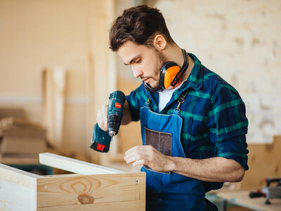 Ein Mann in einem karierten Hemd und einer blauen Schürze benutzt eine elektrische Bohrmaschine, um an einem Holzstück zu arbeiten. Der Mann trägt orange Ohrenschützer um den Hals. Im Hintergrund sind weitere Holzbretter und Werkzeuge zu sehen.