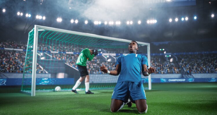Vor einem Fußballtor in einem Stadion sind zwei Fußballspieler in einem grünen und einem blauen Trikot. Der Spieler im grünen Trikot steht im Tor und schlägt die Hände über dem Kopf zusammen. Der Spieler im blauen Trikot kniet und jubelt.