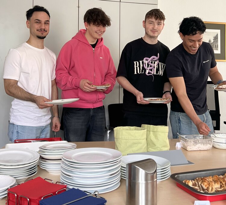 Vier junge Mitarbeiter füllen sich mit einem breiten Grinsen ihre Teller am Buffet mit Brot und Aufstrichen. Auf dem Tisch stehen außerdem mehrere Stapel mit Tellern, Servietten, Brotsäcken, einem Tischabfalleimer und einem Nusszop