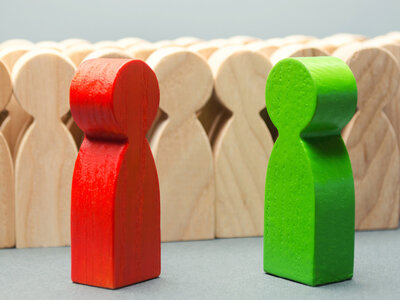 Eine Gruppe von Holzfiguren, wobei zwei Figuren im Vordergrund stehen. Die Figur auf der linken Seite ist rot und die Figur auf der rechten Seite ist grün. Im Hintergrund stehen mehrere neutrale, holzfarbene Figuren.