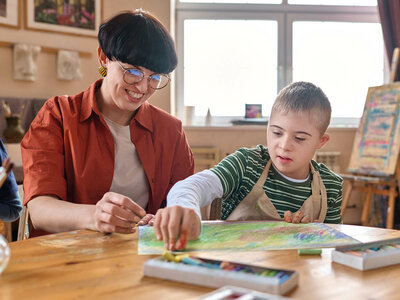 Eine Frau mit kurzen Haaren und Brille sitzt lächelnd an einem Tisch und hilft einem Jungen mit Down-Syndrom beim Malen. Der Junge trägt ein gestreiftes Shirt und eine Schutzschürfe. Beide arbeiten gemeinsam an einem Bild mit bunten Pastellkreiden. Auf dem Tisch liegen weitere Kreiden und eine offene Kreidebox. Im Hintergrund sind Kunstutensilien und ein Fenster zu sehen.