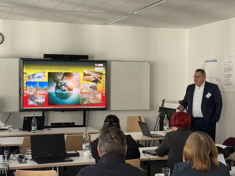 Ein Referent steht vor einer Präsentationsfolie mit bunten Bildern zum Thema Umwelt und Nachhaltigkeit. Teilnehmende sitzen an Tischen, hören zu und machen Notizen.