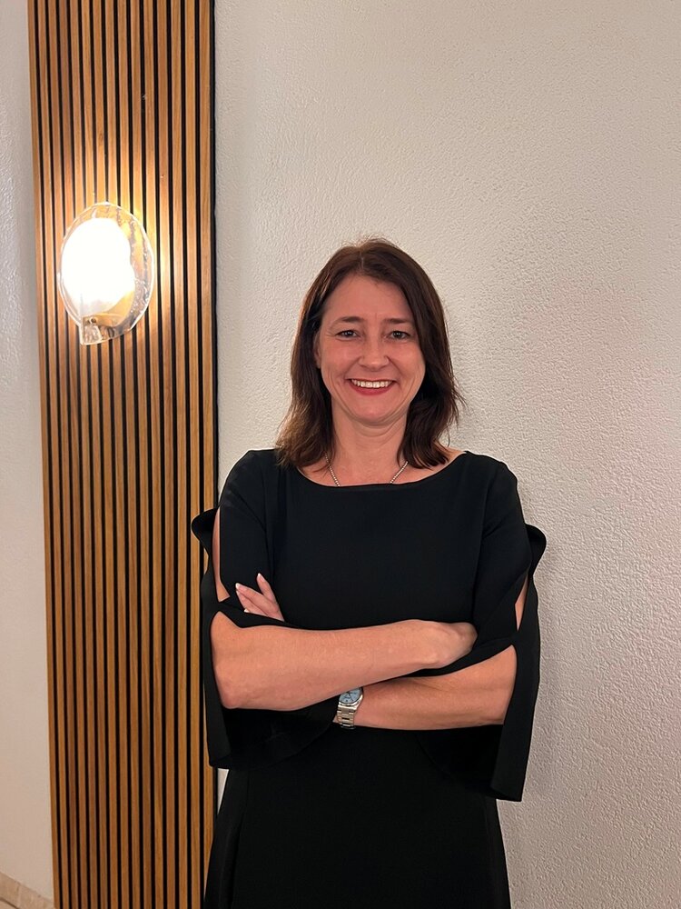 Eine Frau mit schulterlangem dunklem Haar steht mit verschränkten Armen vor einer weißen Wand. Sie trägt ein schwarzes Kleid mit kurzen Ärmeln. Links im Bild befindet sich eine vertikale Holzleiste mit einer runden Wandlampe, die warmes Licht ausstrahlt.