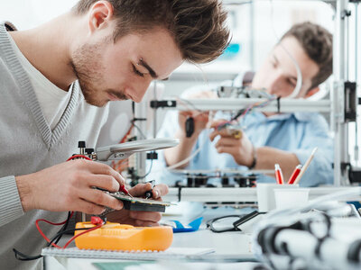 Zwei Personen arbeiten in einem Labor oder einer Werkstatt an elektronischen Komponenten. Die Person im Vordergrund hält eine Leiterplatte und nutzt eine Lupe, während die Person im Hintergrund offenbar mit einem 3D-Drucker oder einem ähnlichen Gerät arbeitet. Auf dem Tisch befinden sich verschiedene Werkzeuge und elektronische Geräte, darunter ein orangefarbenes Multimeter, Kabel sowie Behälter mit Stiften und anderen Kleinteilen.
